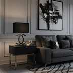 A Clérachic table lamp with a black shade and a dark metal base on a black bedside table, next to a dark grey sofa and a black and white print.