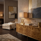 Lit Clérachic gold table lamp with a cream shade on a burr-wood sideboard, beside a decanter and glasses, in a warm, dark-toned room.