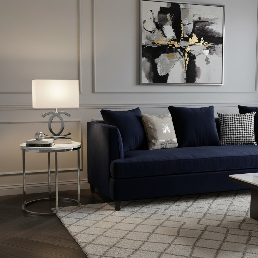 Clérachic table lamp with a white shade and a chrome-toned base on a round side table beside a navy sofa and a black and white print.
