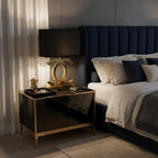 Gold Clérachic table lamp with a black shade on a black and brass bedside table, beside a bed with a navy headboard.