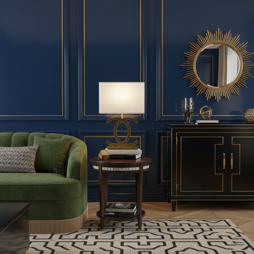 A Clérachic gold table lamp with a cream shade on a round wooden side table, in front of a navy panelled wall with a sunburst mirror.