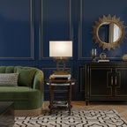 A Clérachic gold table lamp with a cream shade on a round wooden side table, in front of a navy panelled wall with a sunburst mirror.