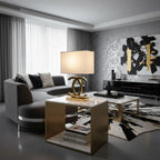 Clérachic gold table lamp with a cream shade on a marble and brass coffee table, in a grey living room with a curved sofa.