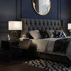 A Clérachic gold table lamp with a cream shade on a mirrored bedside table, beside a bed with a grey tufted headboard and a navy wall.