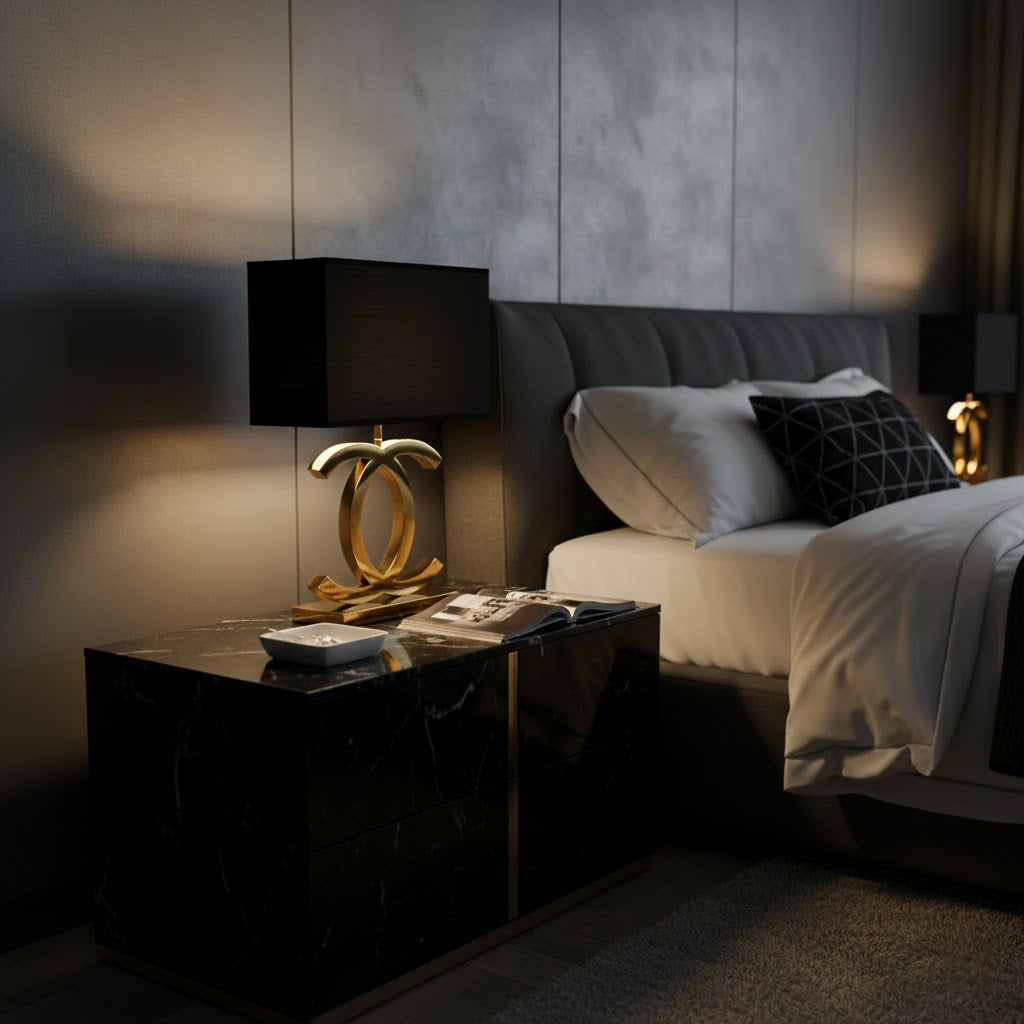 Clérachic gold table lamp with a black shade on a black marble bedside table in a dimly lit bedroom with a grey tufted headboard.