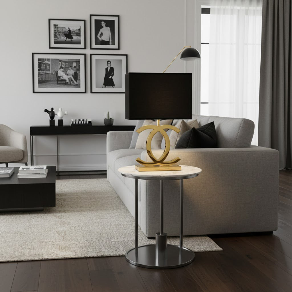 A Clérachic gold table lamp with a black shade on a round side table with a chrome stem, beside a pale grey sofa and framed photographs.
