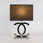 Lit Clérachic chrome table lamp with a black rectangular shade glowing amber, on a polished stainless steel base against a light backdrop.