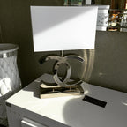 Clérachic chrome table lamp with a white shade on a white console, with a mirrored stand and a vase in the background.
