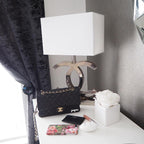 A Clérachic chrome table lamp with a white shade on a white side table, beside a black handbag, a small white flower and a dark curtain.