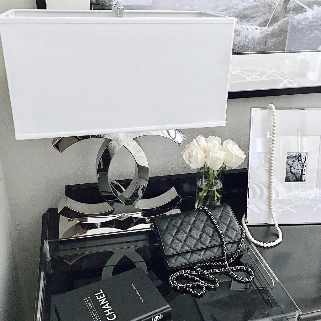Close view of a Clérachic chrome table lamp with a white shade on a dark glossy table, beside white roses, a framed picture and a black handbag.