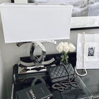 Close view of a Clérachic chrome table lamp with a white shade on a dark glossy table, beside white roses, a framed picture and a black handbag.