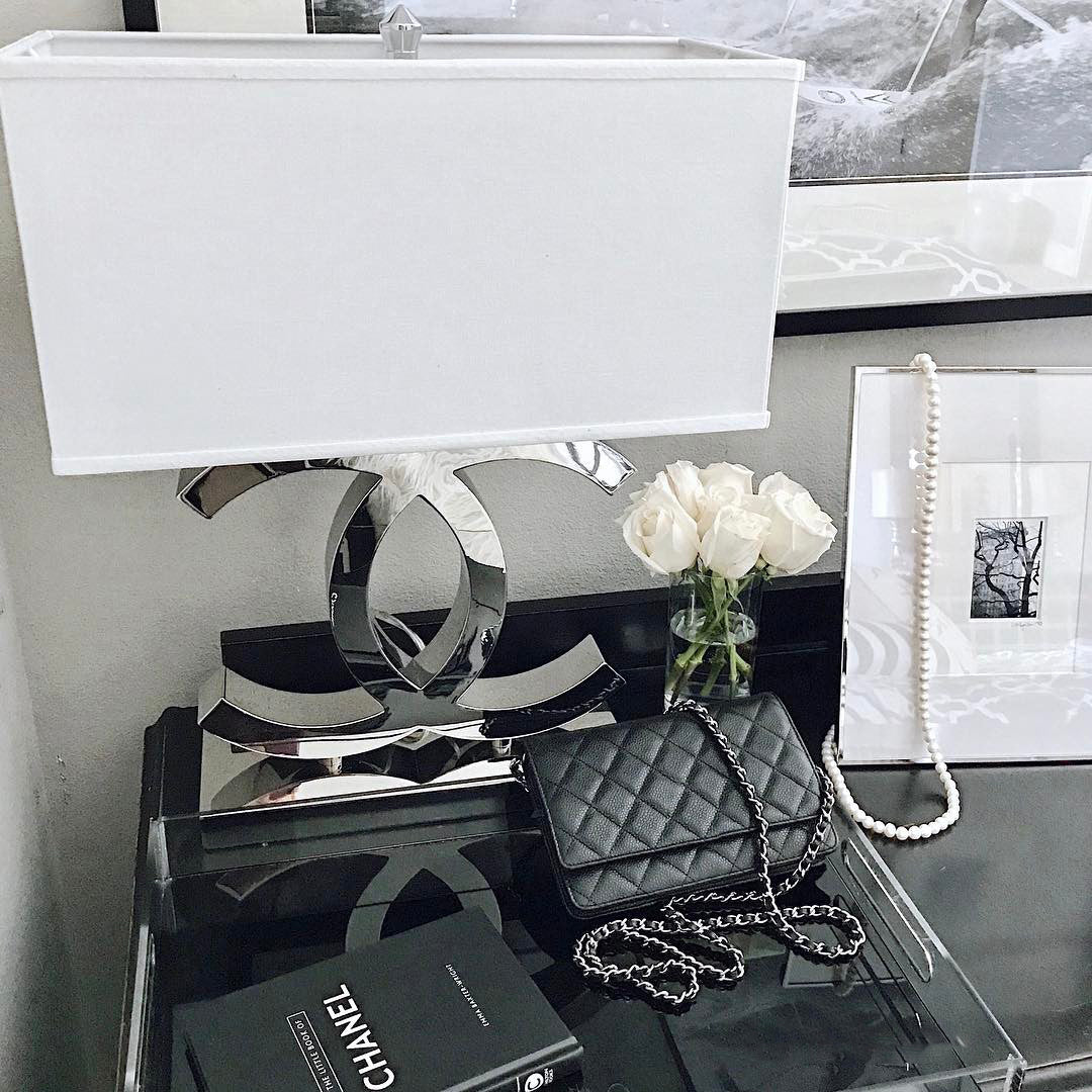 Close view of a Clérachic chrome table lamp with a white shade on a dark glossy table, beside white roses, a framed picture and a black handbag.