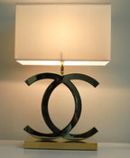 Lit Clérachic table lamp with a white shade glowing warm, its steel loops above a gold base, against a pale green wall.