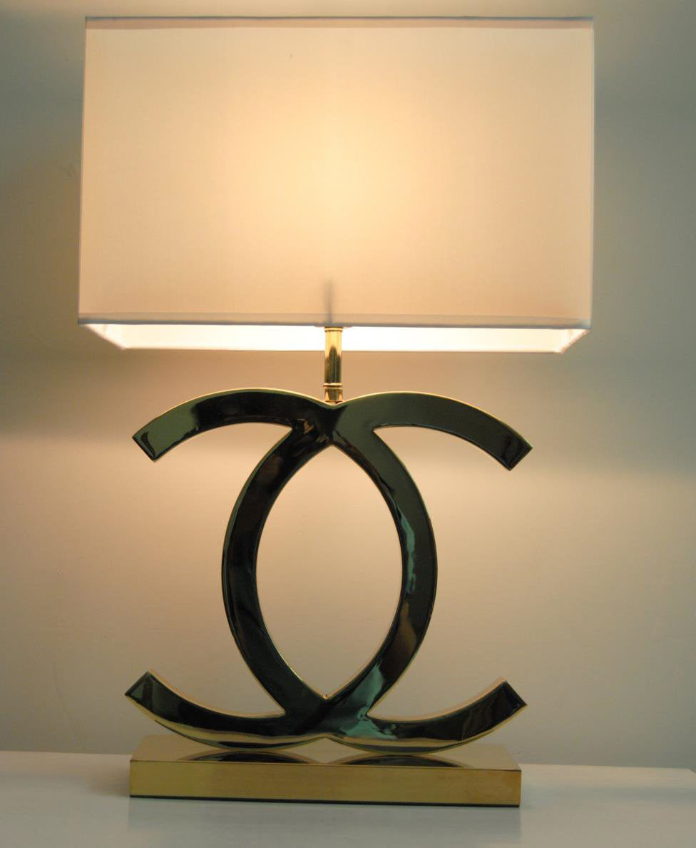 Lit Clérachic table lamp with a white shade glowing warm, its steel loops above a gold base, against a pale green wall.
