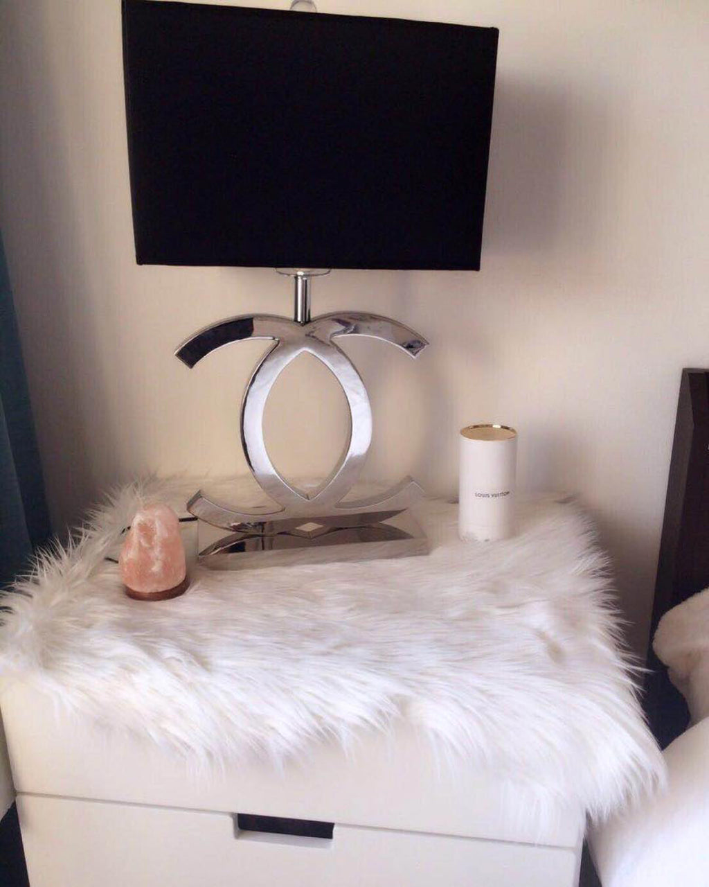 A Clérachic chrome table lamp with a black shade on a white nightstand covered with a fluffy rug, beside a candle and a pink crystal.