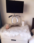 A Clérachic chrome table lamp with a black shade on a white nightstand covered with a fluffy rug, beside a candle and a pink crystal.