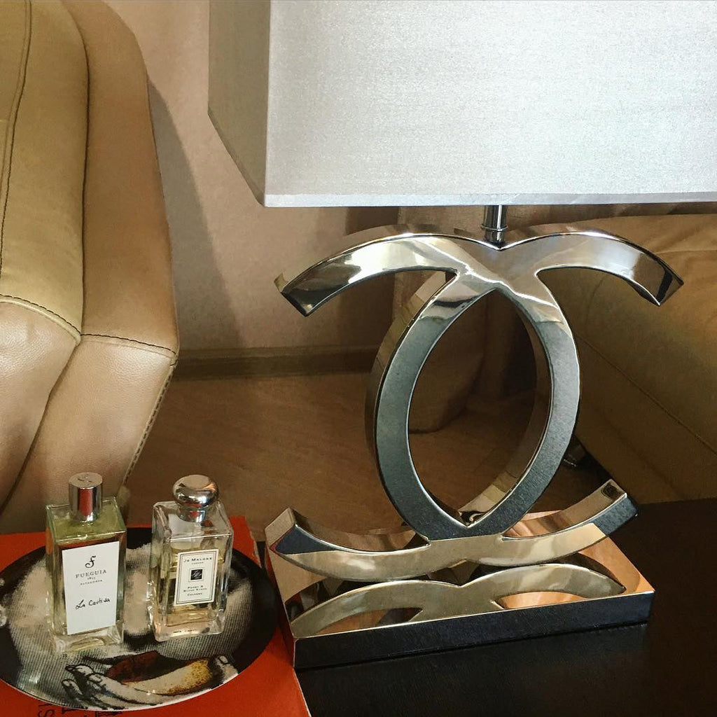 Close view of the polished stainless steel base of a Clérachic table lamp on a dark table, with two perfume bottles on a tray beside it.