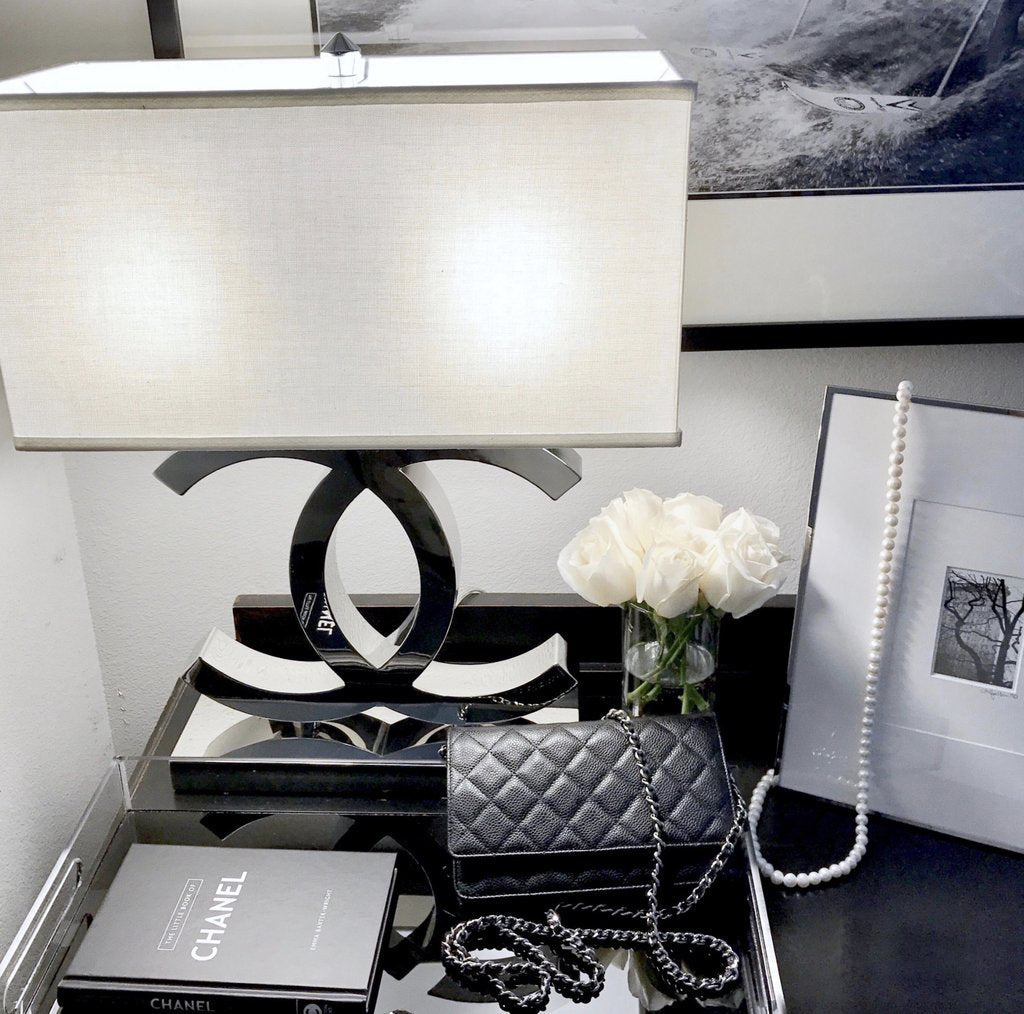 A Clérachic chrome table lamp with a white shade on a mirrored tray table, beside white roses, a black handbag, a framed picture and a book.