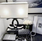 A Clérachic chrome table lamp with a white shade on a mirrored tray table, beside white roses, a black handbag, a framed picture and a book.