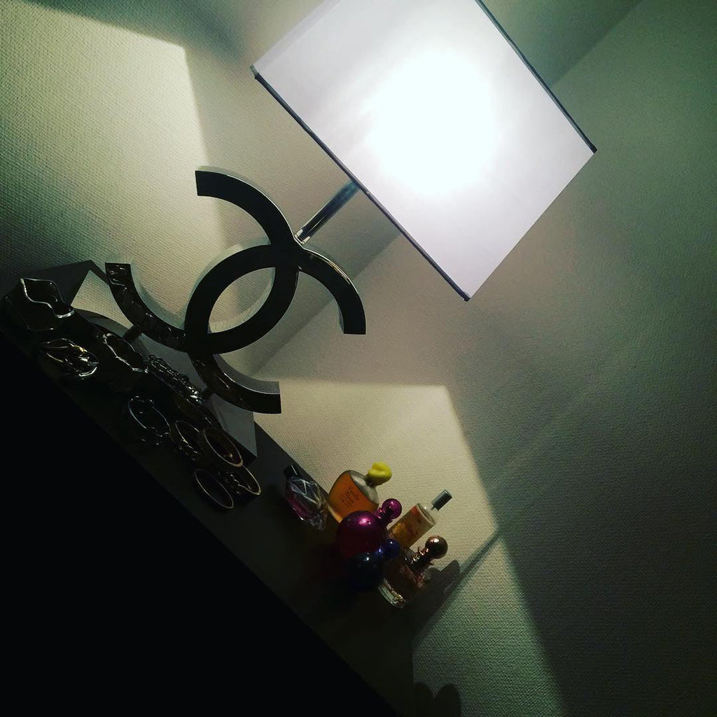 Tilted photo of a Clérachic chrome table lamp with a white shade on a shelf, with small bottles in front and a green wall behind.
