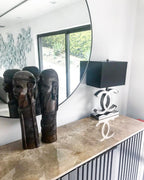 A Clérachic table lamp with a black shade and a polished steel base on a marble console, in front of a round mirror and two dark carved figures.