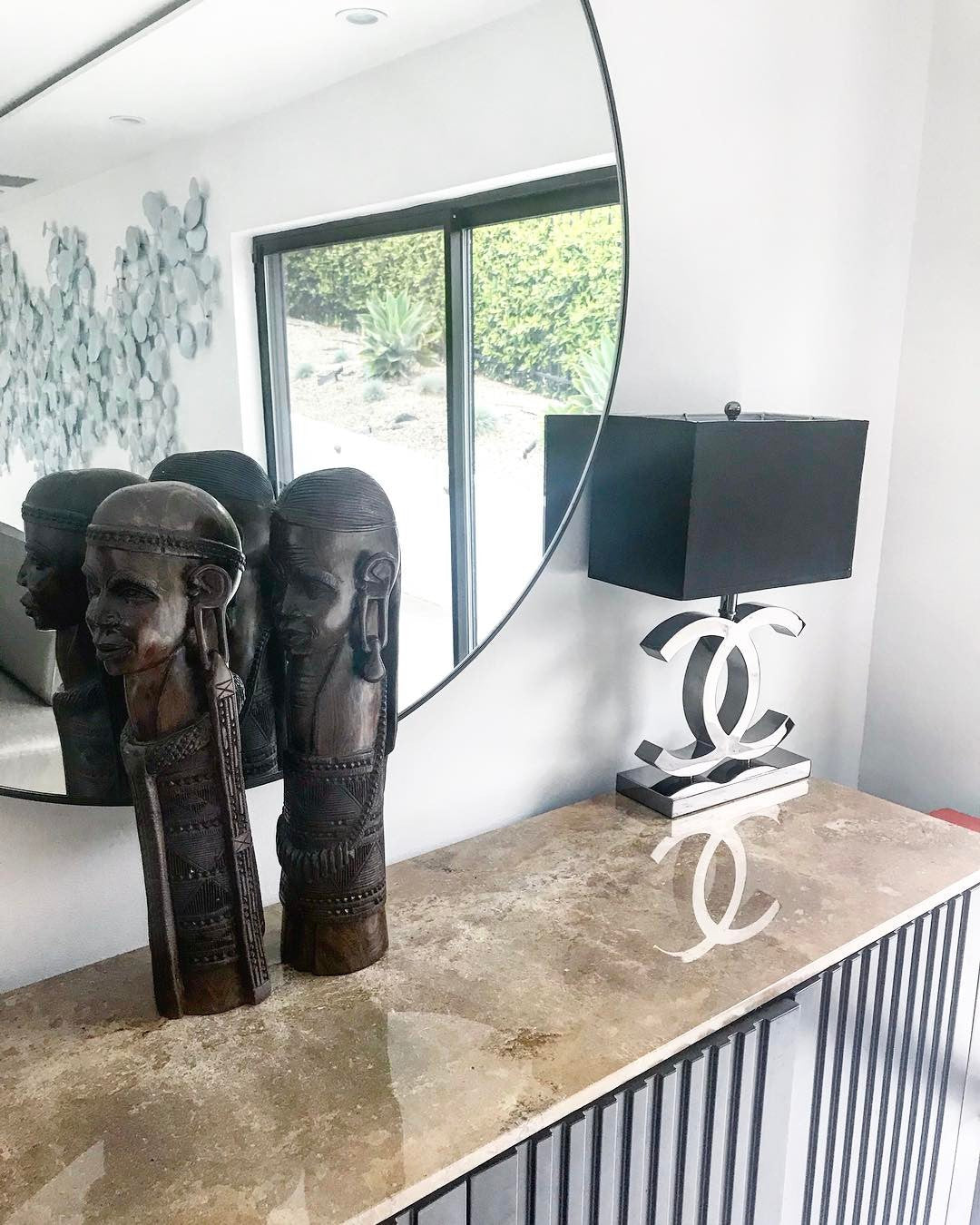 A Clérachic table lamp with a black shade and a polished steel base on a marble console, in front of a round mirror and two dark carved figures.