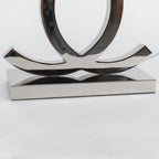 Close view of the lower curve of the loops and the stepped steel base on this Clérachic table lamp, against a pale backdrop.