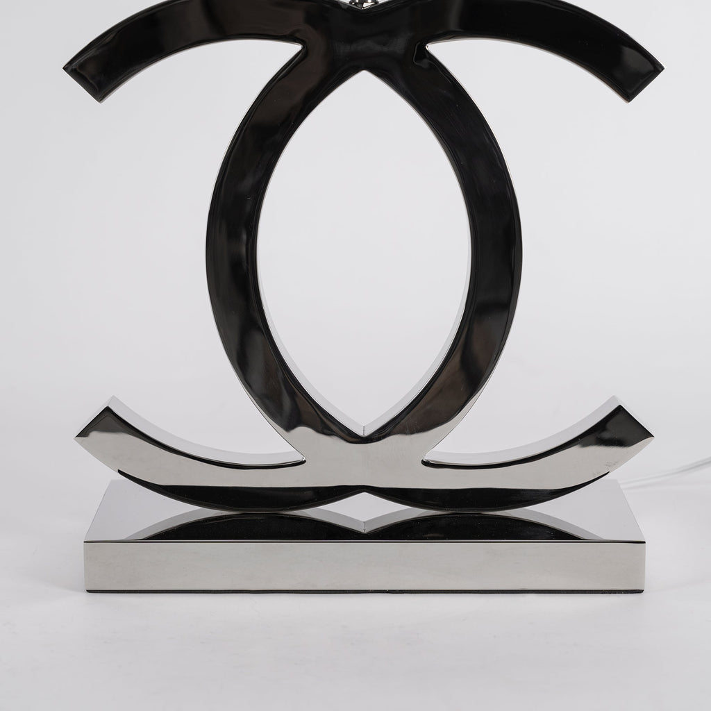 Close view of the polished dark chrome loops and the flat rectangular base of this Clérachic table lamp, against a pale backdrop.