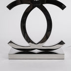 Close view of the polished dark chrome loops and the flat rectangular base of this Clérachic table lamp, against a pale backdrop.