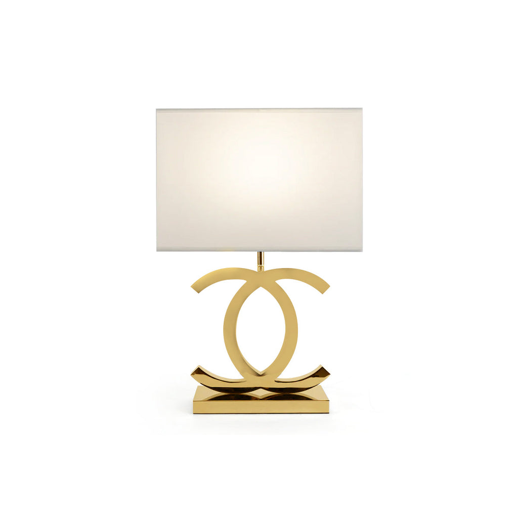 A Clérachic gold table lamp with a white rectangular shade, its polished gold loops on a gold base, on a white backdrop.
