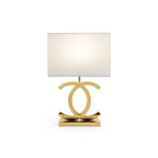 A Clérachic gold table lamp with a white rectangular shade, its polished gold loops on a gold base, on a white backdrop.