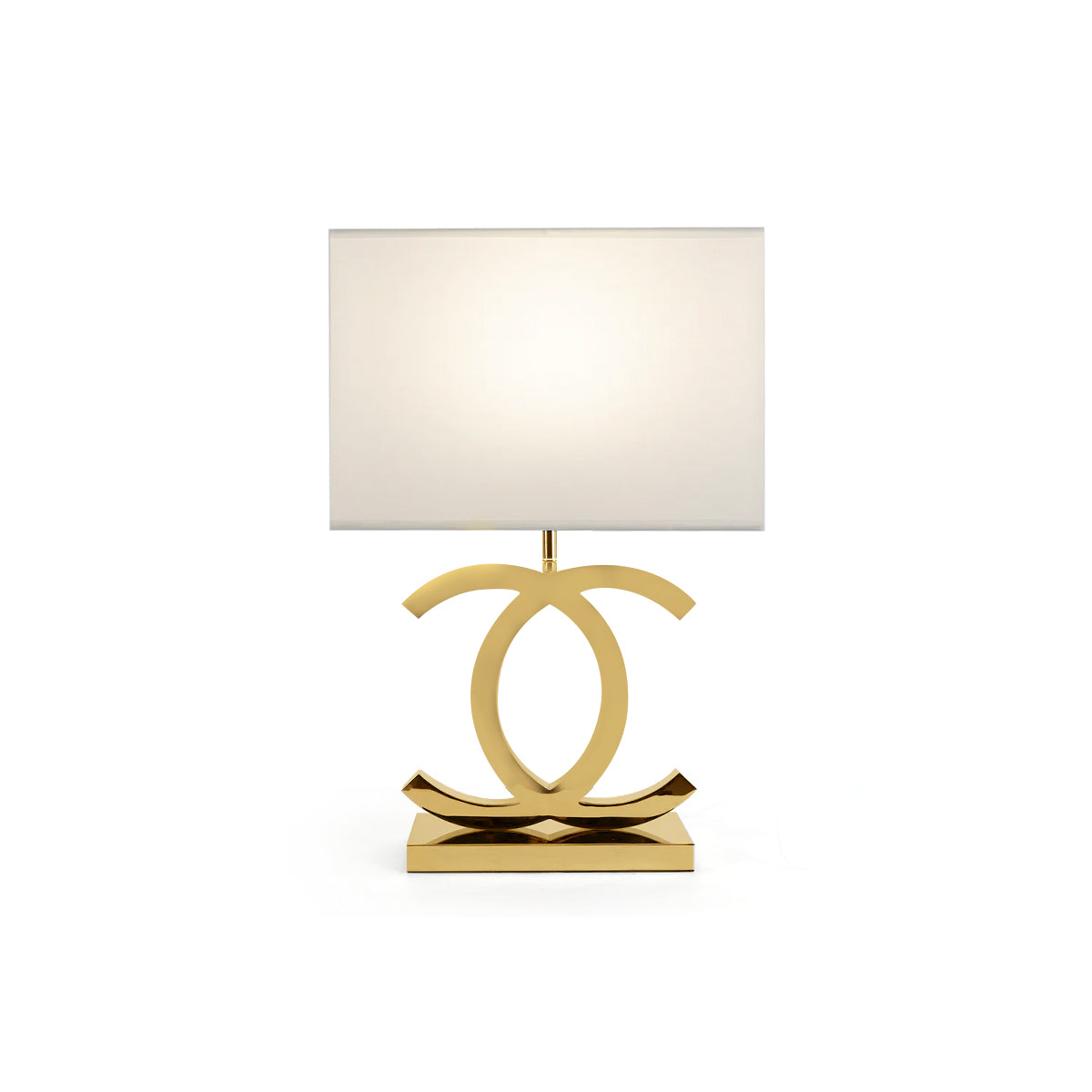 A Clérachic gold table lamp with a white rectangular shade, its polished gold loops on a gold base, on a white backdrop.