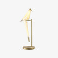 A Clérachic table lamp shaped like a folded paper bird, perched on a slim gold stand with a round gold base, on a plain white backdrop.