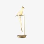 A Clérachic table lamp shaped like a folded paper bird, perched on a slim gold stand with a round gold base, on a plain white backdrop.