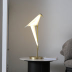 The glowing white folded-paper bird of this Clérachic table lamp, on a gold stand over a round dark table with a book, against a grey wall.