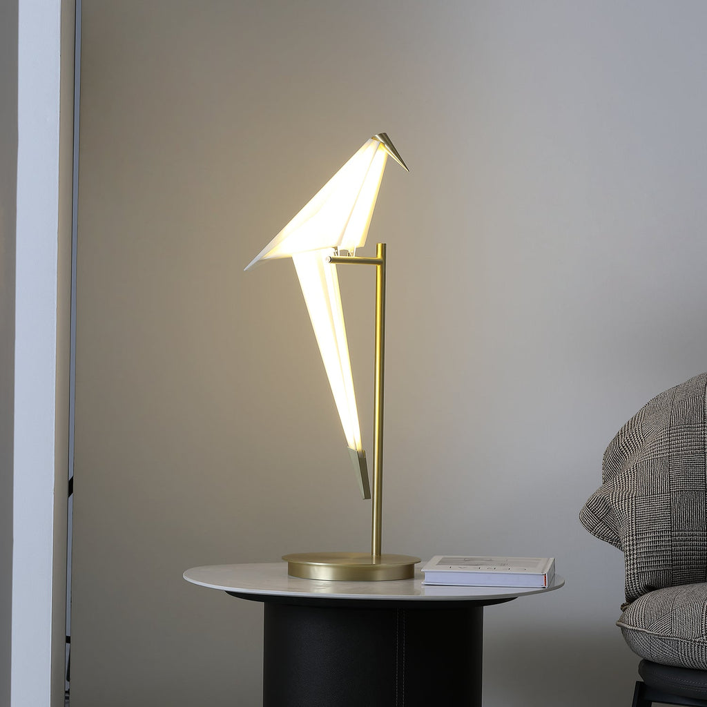 Warm light inside the paper bird of this Clérachic lamp, on a gold stand on a marble-topped table beside a grey tweed armchair.