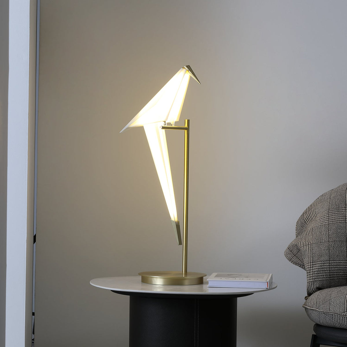 Warm light inside the paper bird of this Clérachic lamp, on a gold stand on a marble-topped table beside a grey tweed armchair.