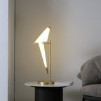 Seen from the left, the lit bird on this Clérachic table lamp leans over its gold stand, with a book on the table beside the base.