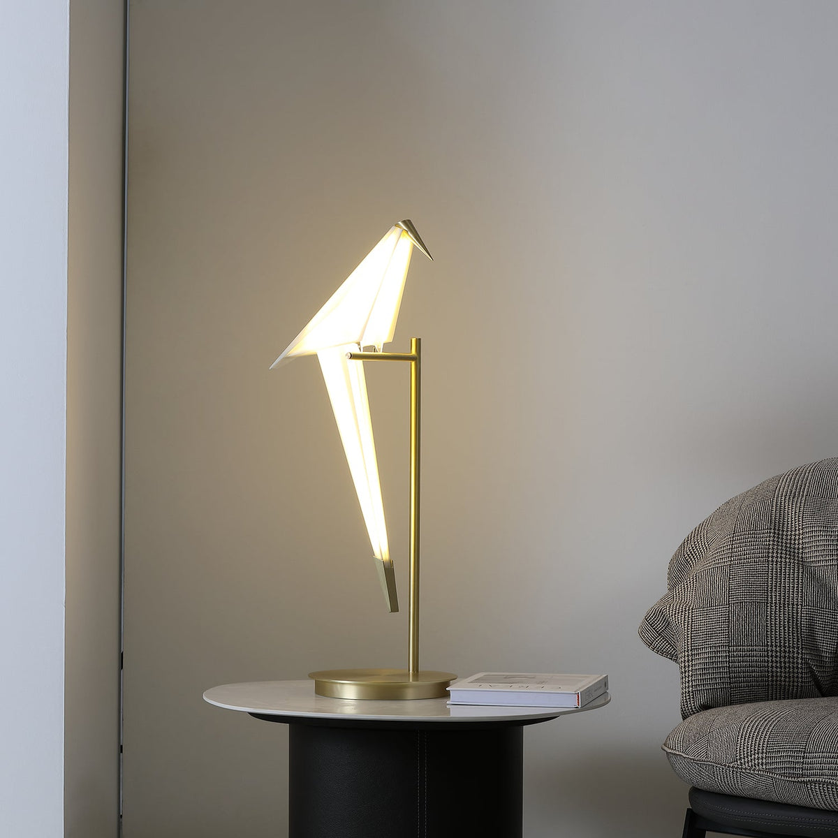 Seen from the left, the lit bird on this Clérachic table lamp leans over its gold stand, with a book on the table beside the base.