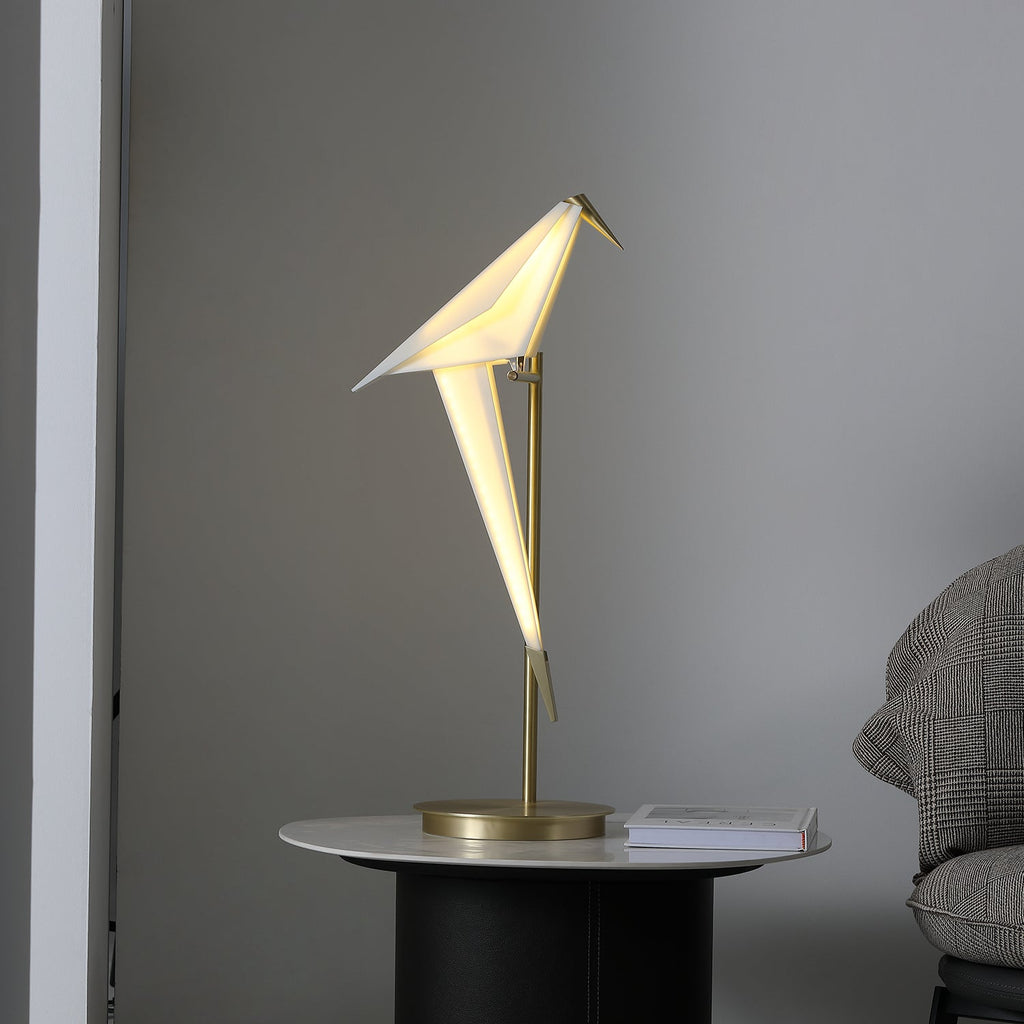 Straight-on view of a lit Clérachic bird lamp, its folded white body tilted on a slim gold stem above a round gold base.