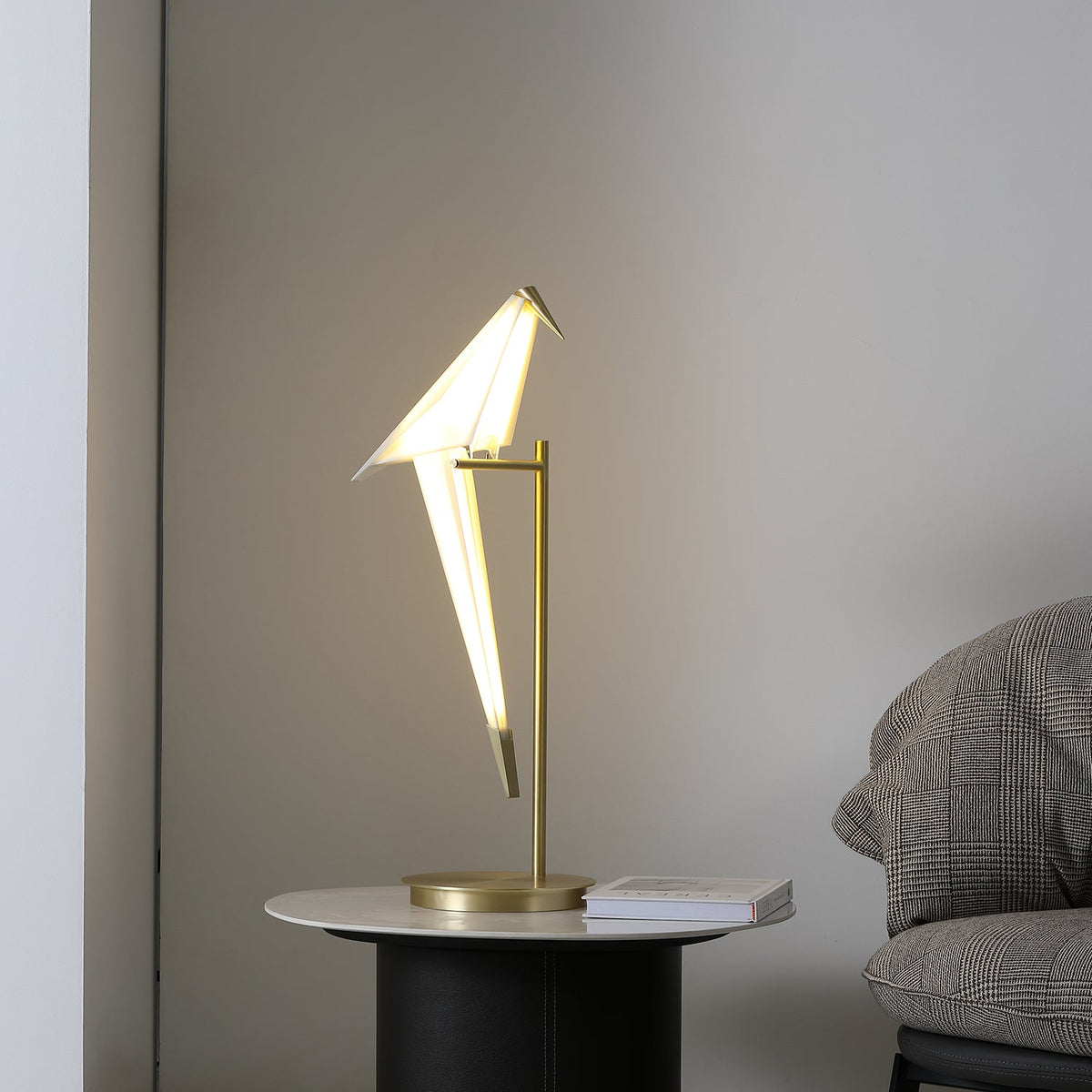 The bird of this Clérachic table lamp, lit and tilted forward on a gold stand, with a stack of books on the round table.