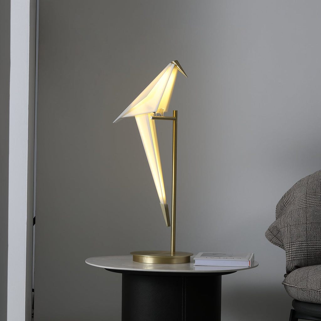 Close view of the lit paper bird of this Clérachic table lamp, with its gold stand and base on a round table by a grey armchair.