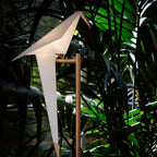 A lit white paper bird on a gold stand, seen among dark green palm leaves, on this Clérachic table lamp.