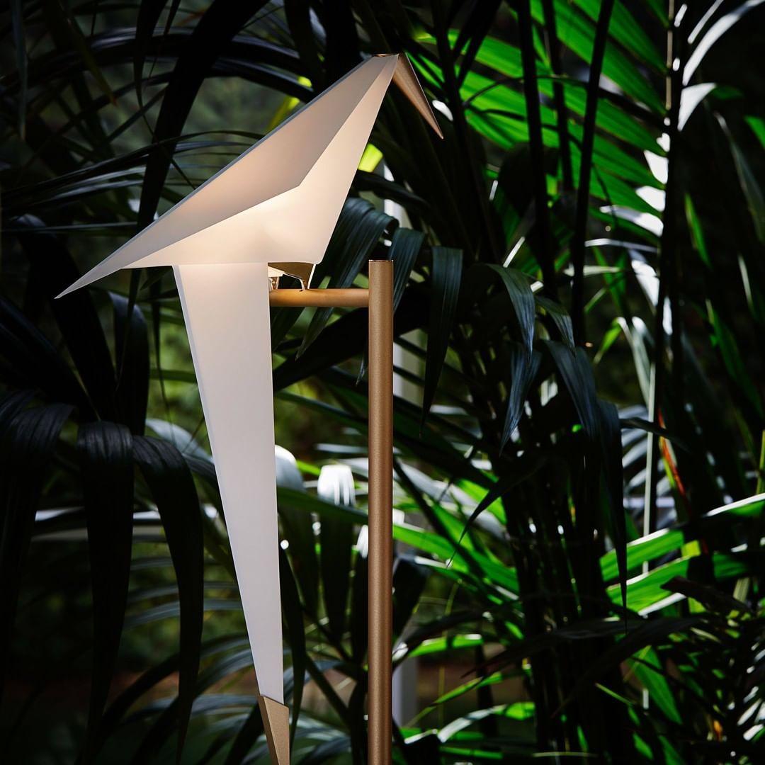 A lit white paper bird on a gold stand, seen among dark green palm leaves, on this Clérachic table lamp.