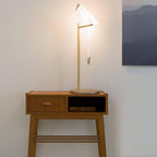 A lit Clérachic bird lamp on a gold stand, set on a teak side table with a drawer, in front of a pale wall and a mountain print.