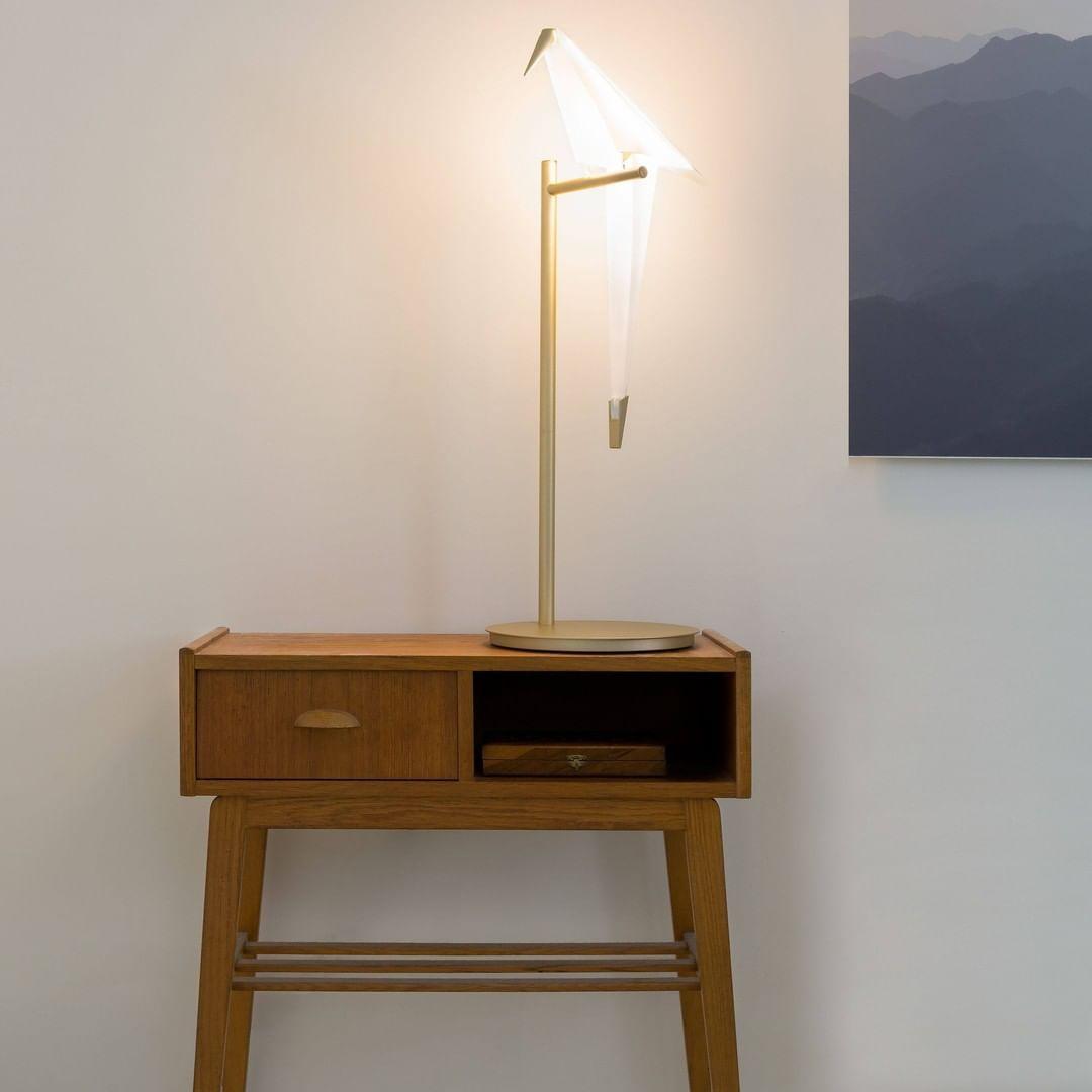 A lit Clérachic bird lamp on a gold stand, set on a teak side table with a drawer, in front of a pale wall and a mountain print.