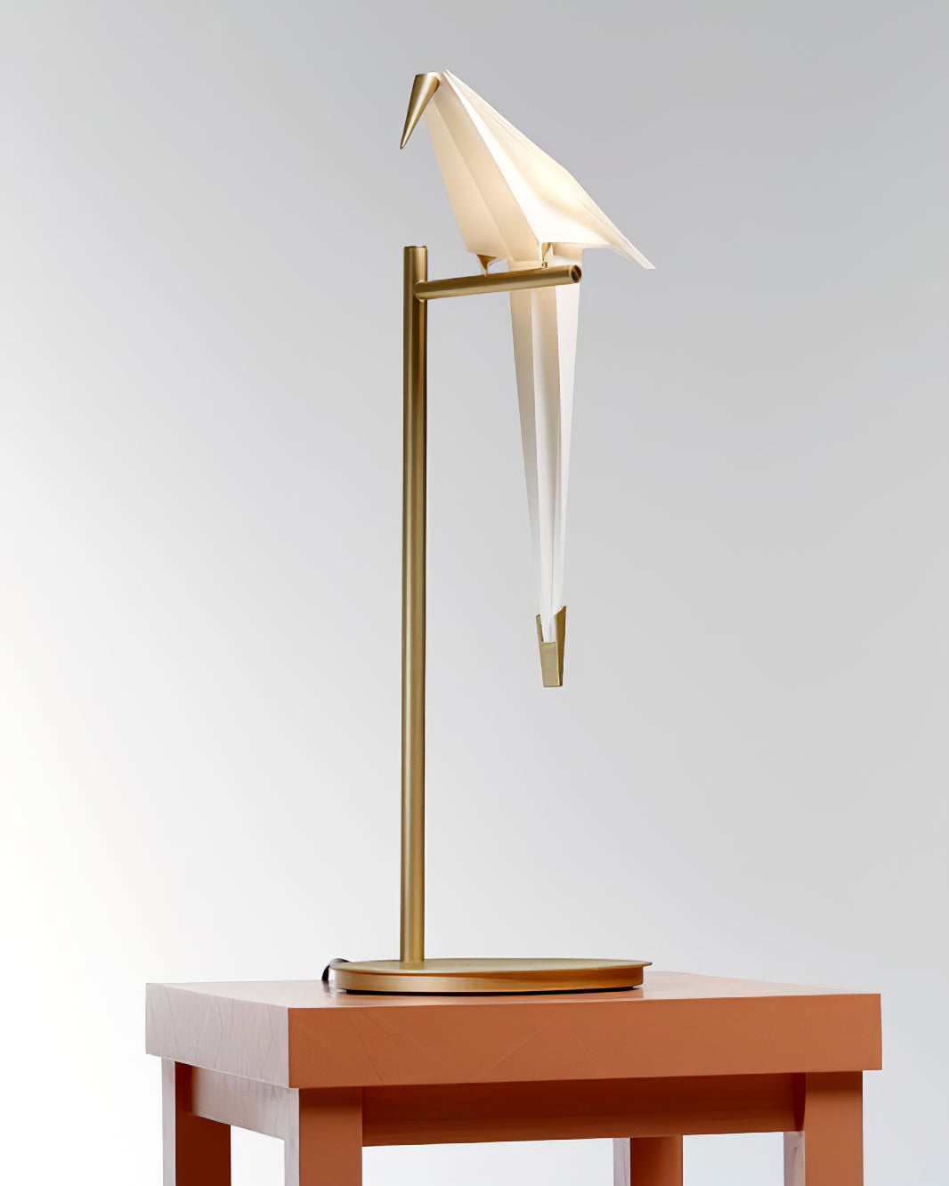 A Clérachic bird table lamp on a gold stand and thin round base, on a terracotta-red table against a pale backdrop.