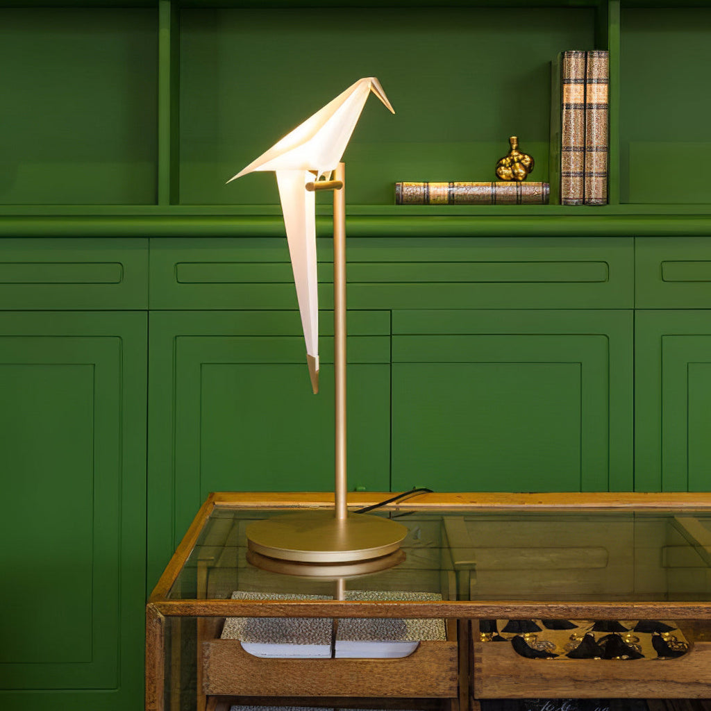 A Clérachic table lamp with a folded white bird on a gold stand, on a glass-topped table in front of a dark green cabinet.