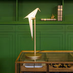 A Clérachic table lamp with a folded white bird on a gold stand, on a glass-topped table in front of a dark green cabinet.