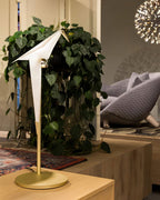 The white paper bird of this Clérachic lamp on a gold stand, with a trailing green plant behind it and a grey chair beyond.